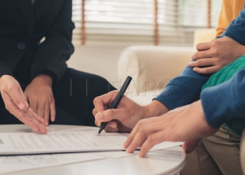 Happy young Asian couple and realtor agent. Cheerful young man signing some documents while sitting at desk together with his wife. Buying new house real estate. Signing good condition contract.