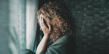 Sad and worried woman side portrait at home in the dark looking outside the window. Worried and lonely female alone indoor. Concept of end of relationship and unsuccessful emotion. Lady touching face