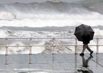 person protecting with umbrella in rainy and windy day walking on promenade with rough sea