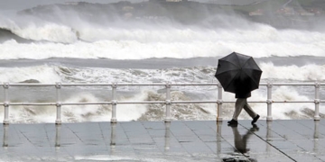 person protecting with umbrella in rainy and windy day walking on promenade with rough sea