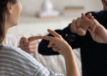 Young positive couple sit on sofa at home communicating using sign language, friends talking indoor showing fingers gestures, having fun, leading pleasant conversation, people with disability concept