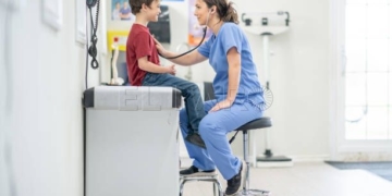 A sweet young boy sits up on an exam table as his female nurse takes a close listen to his heart with her stethoscope.  He is dressed casually and sitting still as she listens closely.