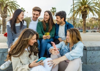 Happy group of student people with mobile phones having fun sitting outdoors. Millennial generation diverse friends laughing enjoying time together social gathering in city street. Youth lifestyle.